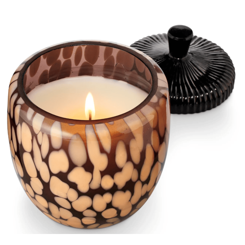 A lit candle in a round glass jar with a brown and beige spotted pattern, shown with its black ribbed lid on a white background.