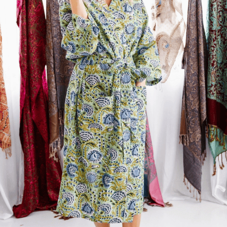 A woman modeling a long-sleeved, belted kaftan dress with a green, blue, and yellow floral block print, standing in front of a white studio background with hanging scarves.