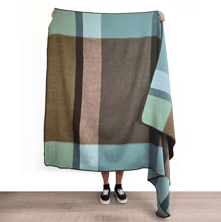 A person holds up a large woven blanket with a large-scale geometric block pattern in muted earthy tones of brown, sage green, beige, and slate blue, standing on a light wood floor against a white wall.