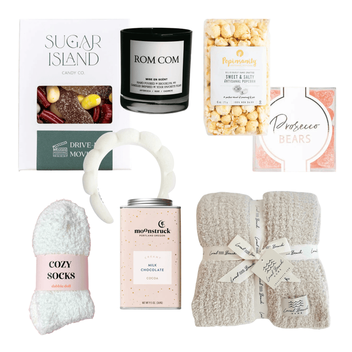 A curated gift basket or bundle containing multiple items: a Sugar Island candy box, a Rom Com candle, a jar of peppermint popcorn, a Prosciore Bears soap bar, a white curved accessory, a pink-labeled cozy sock, a Mochi Chocolate tin, and a folded cream towel with a ribbon. All items are arranged on a plain white background.