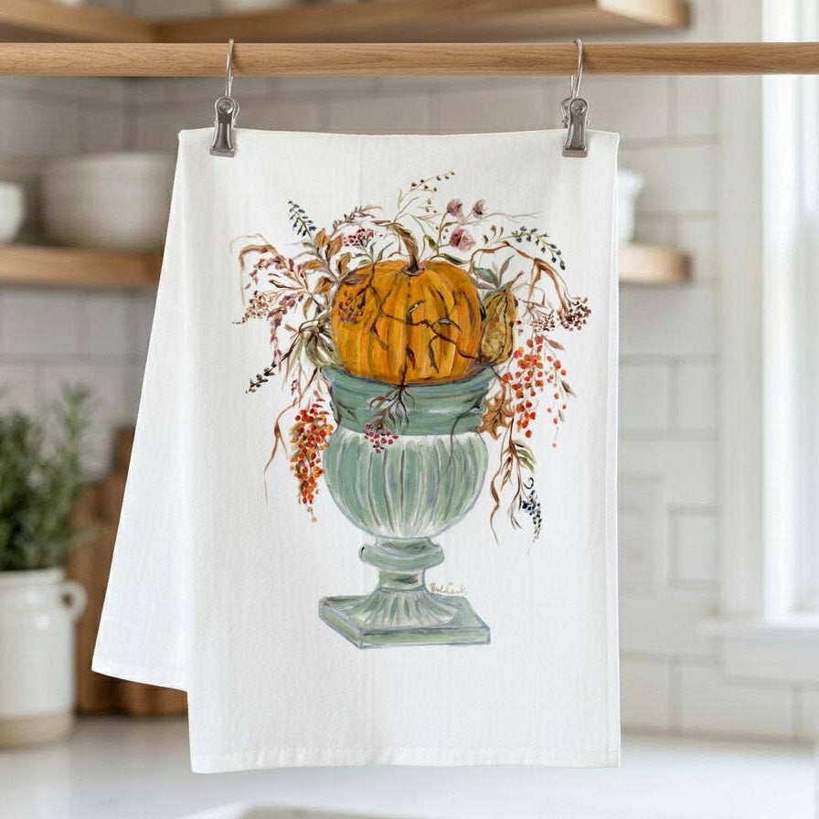 A white kitchen tea towel hanging from a wooden rod with metal clips, featuring a printed illustration of an orange pumpkin in a pale green urn with autumn foliage. The towel is photographed in a bright, softly blurred kitchen setting.