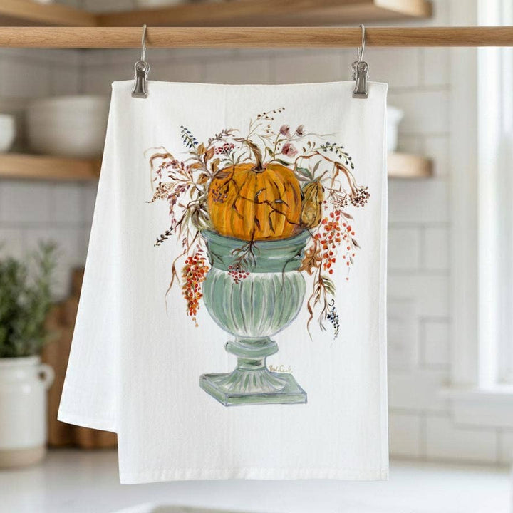 A white kitchen tea towel hanging from a wooden rod with metal clips, featuring a printed illustration of an orange pumpkin in a pale green urn with autumn foliage. The towel is photographed in a bright, softly blurred kitchen setting.