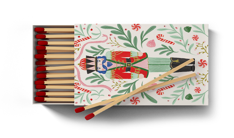 A decorative matchbox filled with wooden matches and red tips, shown open on a dark green background. The matchbox lid features a festive nutcracker illustration with candy cane and holiday motifs.