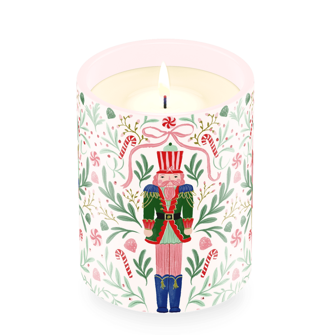 A lit jar candle with a white wax body and a printed holiday label featuring a nutcracker surrounded by green leaves, pink bows, candy canes, and small pink hearts on a white background.
