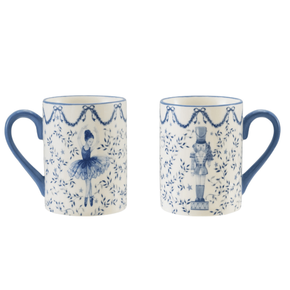 Two white ceramic mugs with blue handles and blue printed illustrations, shown side by side on a white background. One mug features a ballerina under a garland pattern, and the other features a nutcracker figure.
