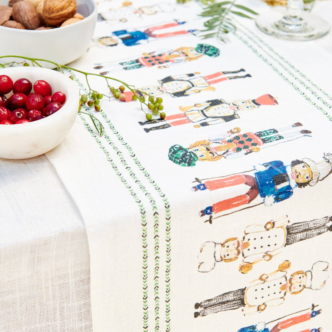 A white table runner with a colorful watercolor print of nutcrackers and decorative green borders, styled on a dining table with bowls of cranberries and walnuts.