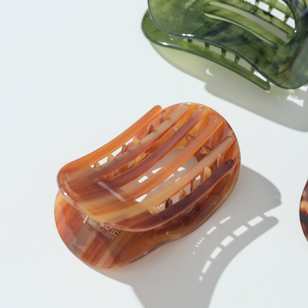 A translucent tortoiseshell hair claw clip in warm amber-brown tones, photographed on a clean white surface with soft shadows. Another green clip is partially visible in the upper right, and part of another clip appears at the lower right edge.