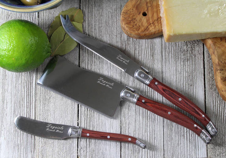 Three Laguiole cheese knives with reddish-brown wood handles and silver blades, arranged on a rustic white-washed wooden table alongside a lime, bay leaf, cheese, and olive wood cutting board.