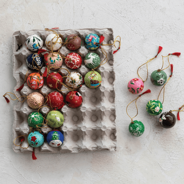 A set of colorful hand-painted Christmas egg ornaments displayed in a brown cardboard egg carton, with several individual ornaments arranged to the right on a light textured background.