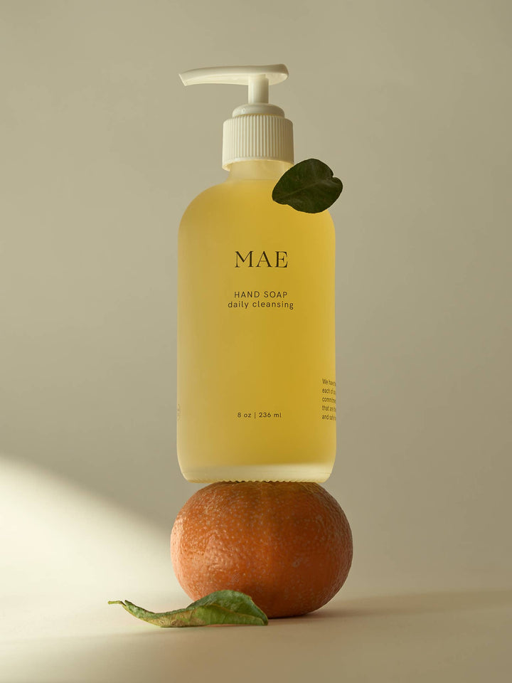 Yellow hand soap bottle labeled 'MAE' on a beige background with an orange and leaf.