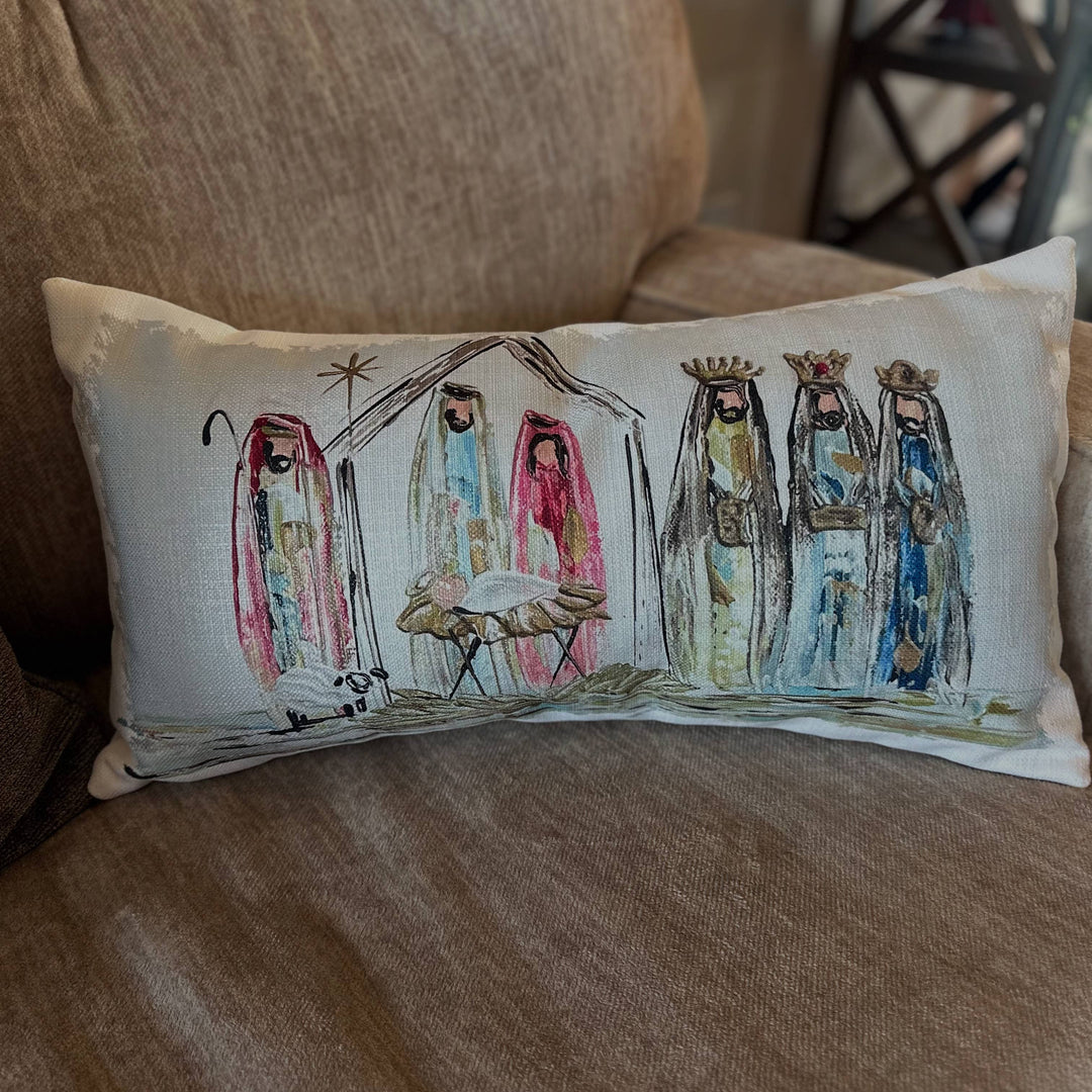 A rectangular throw pillow with a hand-painted nativity scene, resting on a textured brown fabric couch.