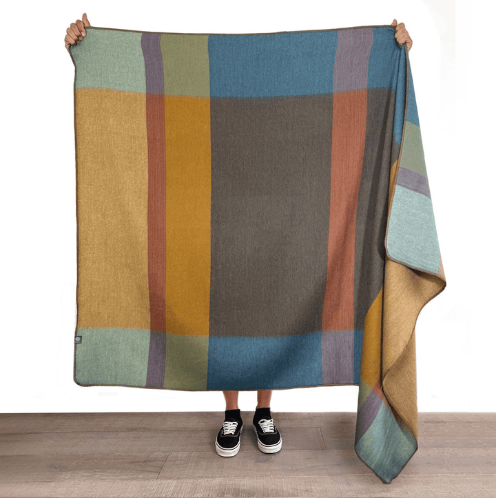 A person holds up a large woven blanket with a multicolor plaid pattern, showing its texture and full design. The blanket features blocks of mustard yellow, slate blue, taupe, sage green, and rust orange.