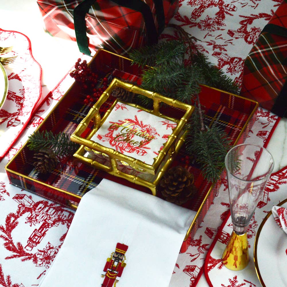 A styled holiday table setting featuring a red and green plaid tray with bamboo-style gold trim, a matching napkin with a nutcracker print, pinecones, greenery, and red-and-white patterned table linens.