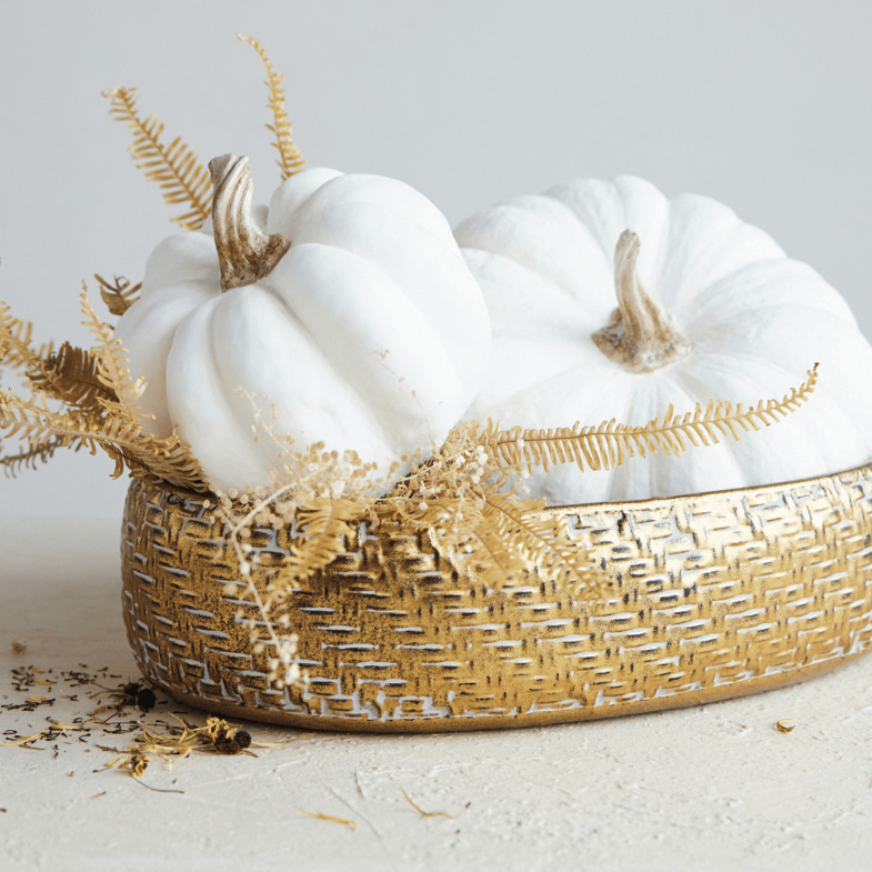 Two small white decorative pumpkins arranged in a woven gold basket with dried gold ferns, photographed on a light neutral surface against a pale gray background.