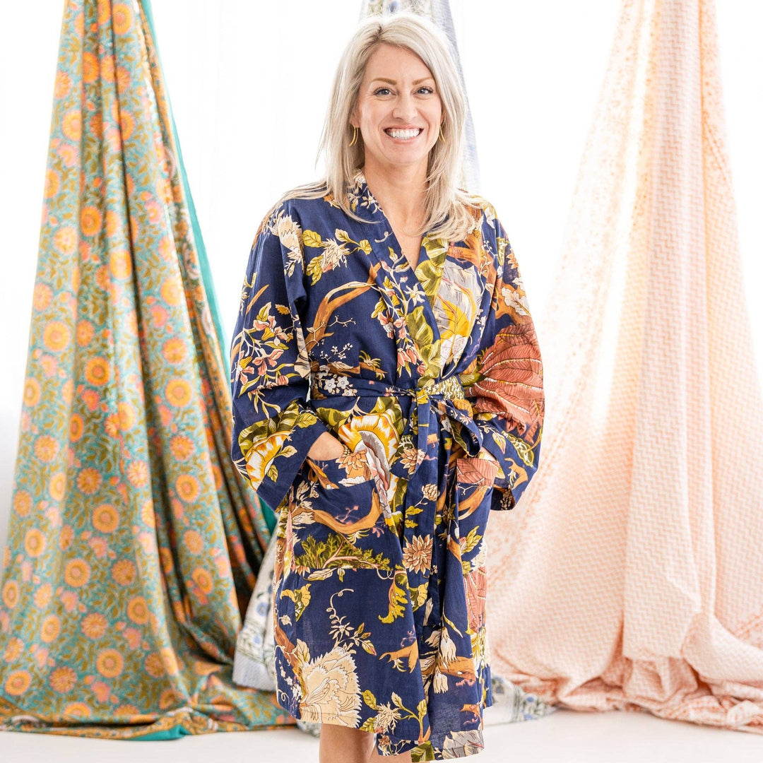 A smiling blonde woman stands indoors wearing a navy kimono-style robe with a multicolor floral and bird print. Draped fabric panels in teal-orange and blush pink hang behind her against a bright white background.