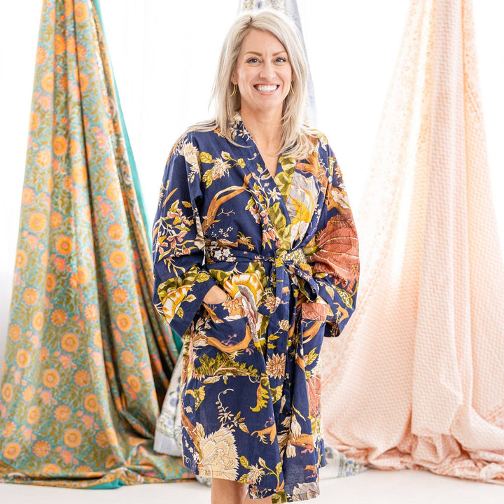 A smiling blonde woman stands indoors wearing a navy kimono-style robe with a multicolor floral and bird print. Draped fabric panels in teal-orange and blush pink hang behind her against a bright white background.