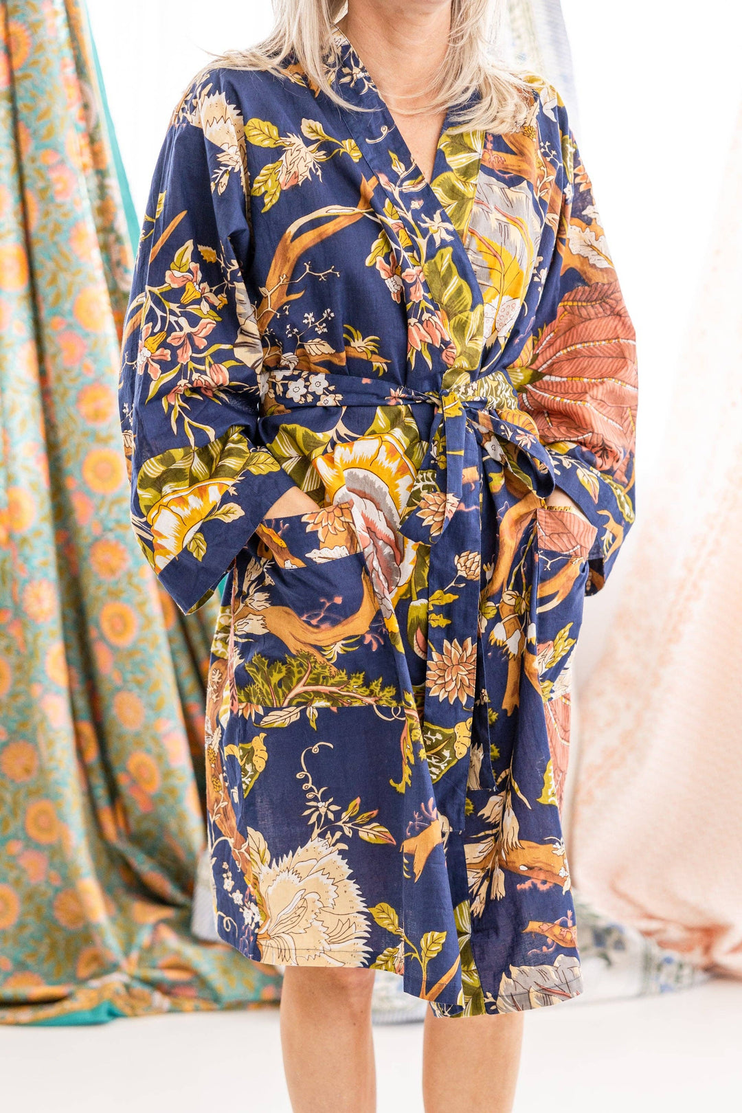 A woman wearing a navy blue kimono-style robe with a colorful botanical and animal print, featuring foxes, birds, and floral motifs in yellow, orange, green, and white. The robe has a tie waist and side pockets, shown against a soft, draped fabric backdrop.