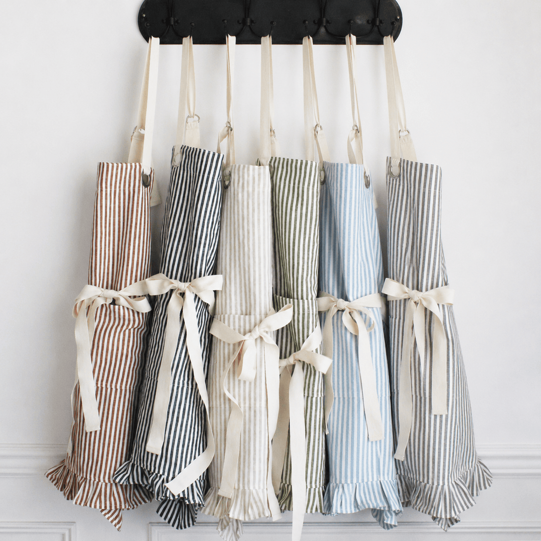Six striped kitchen aprons in different colors hang from a black tufted wall rack against a white wall. The aprons have cream ties and ruffled hems, and are shown in pinkish-brown, navy, cream, sage green, light blue, and gray stripes.