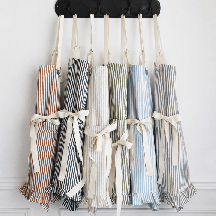 Six striped kitchen aprons in different colors hang from a black tufted wall rack against a white wall. The aprons have cream ties and ruffled hems, and are shown in pinkish-brown, navy, cream, sage green, light blue, and gray stripes.