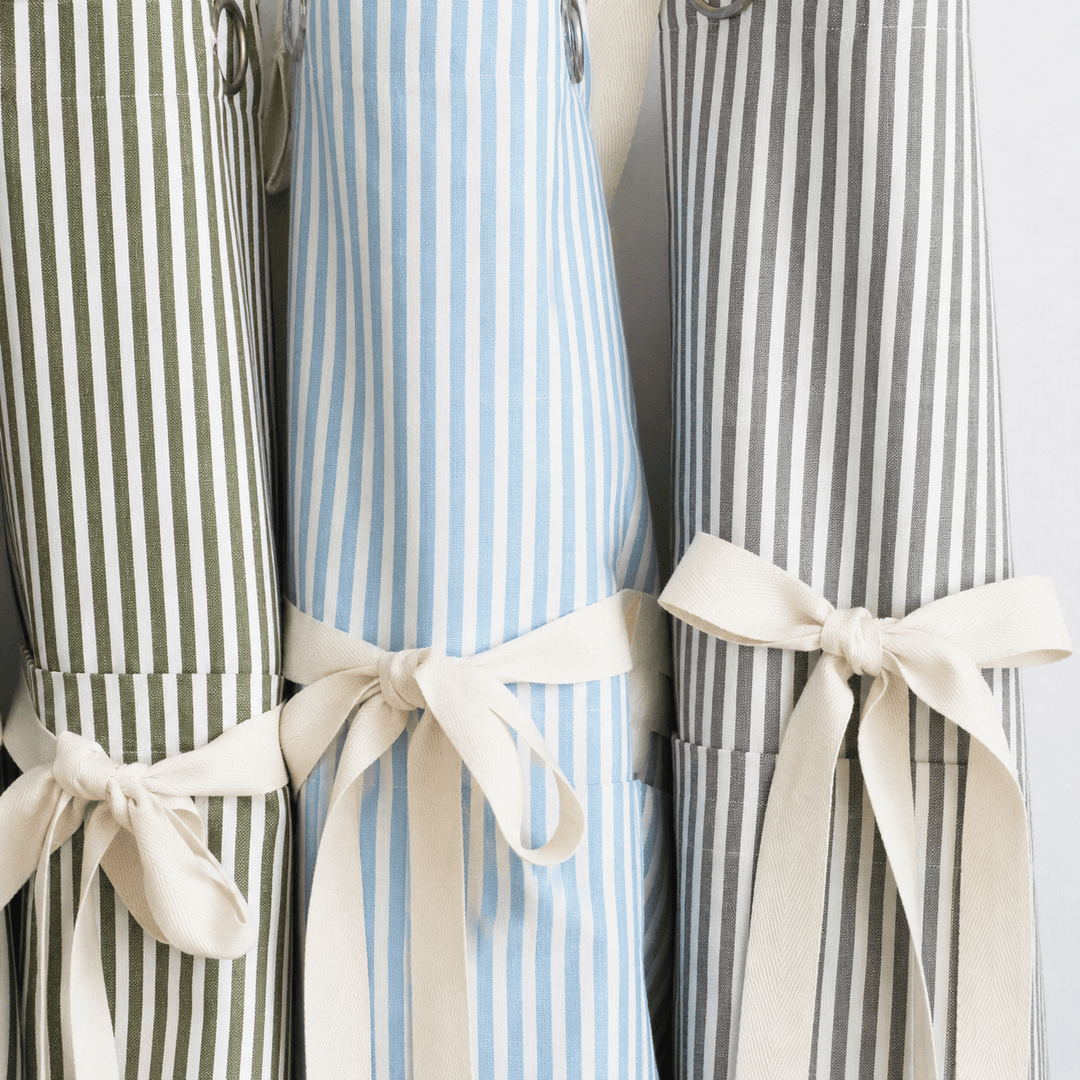 Three striped kitchen aprons tied with cream ribbon bows, shown side by side against a light background. The left apron has olive green and white stripes, the center apron has light blue and white stripes, and the right apron has gray and white stripes.