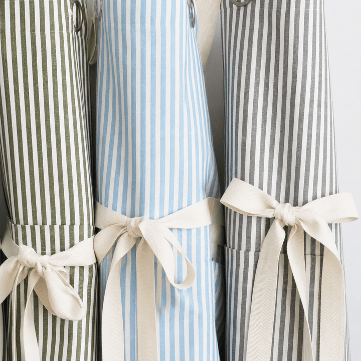 Three striped kitchen aprons tied with cream ribbon bows, shown side by side against a light background. The left apron has olive green and white stripes, the center apron has light blue and white stripes, and the right apron has gray and white stripes.