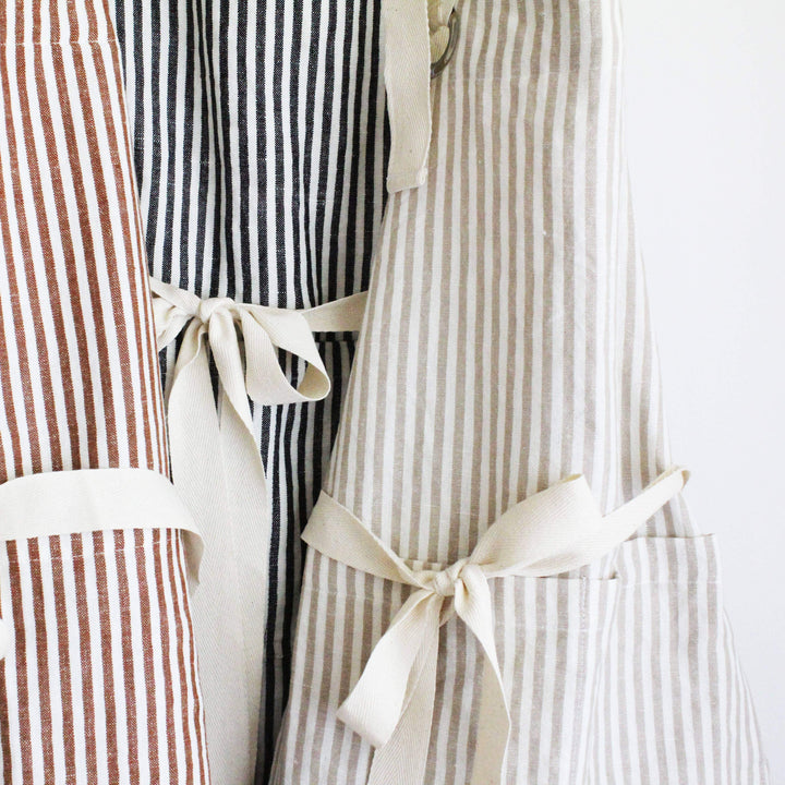 Three striped aprons hanging side by side against a plain white background. The left apron has vertical brown and white stripes, the middle apron has vertical black and white stripes, and the right apron has vertical beige and white stripes with a cream fabric tie belt and front pocket.