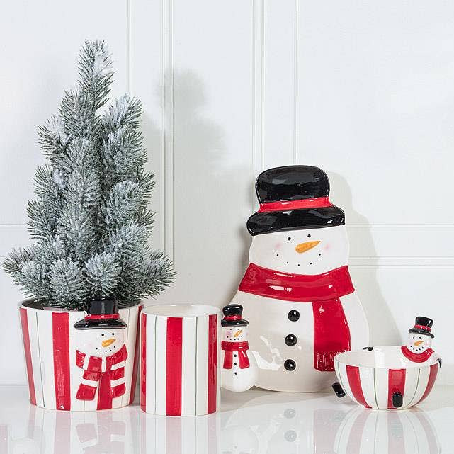 A set of ceramic holiday snowman decorations including a small artificial frosted tree, two striped planters, a larger snowman figure, a small snowman figure, and a striped bowl, arranged on a white surface against a white paneled wall.