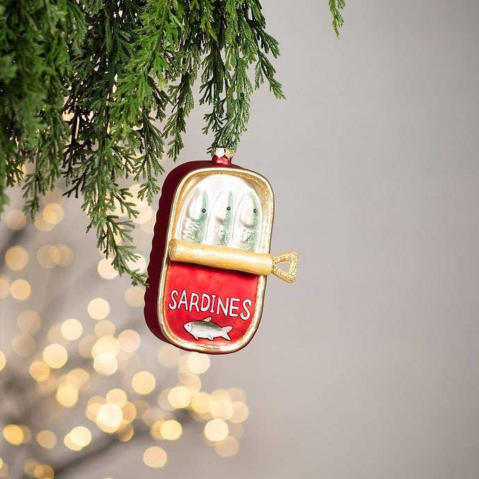 A Christmas ornament shaped like an open can of sardines hanging from green pine branches. The ornament is red with gold trim, white lettering that reads 'SARDINES,' and silver fish illustrations on the inside.