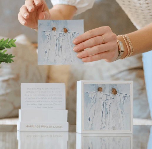 Person holding a prayer card with artistic design, surrounded by similar cards and a plant.