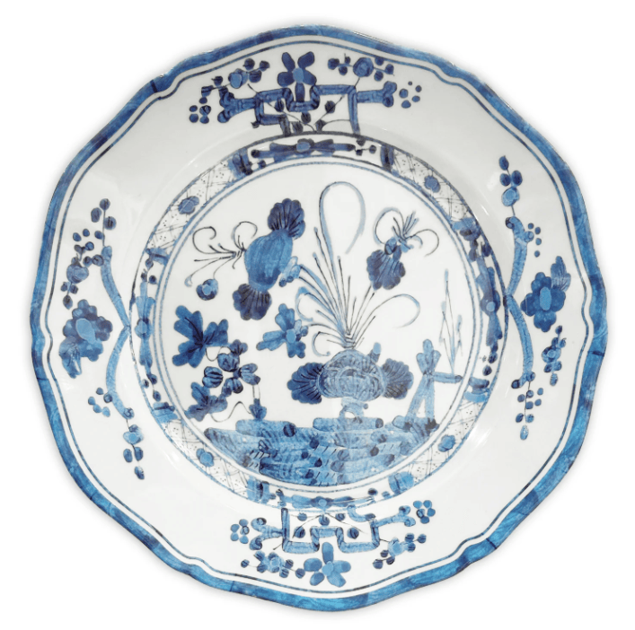 A round white melamine plate with a scalloped rim, decorated with blue floral and botanical patterns and a central landscape motif, shown against a plain white background.