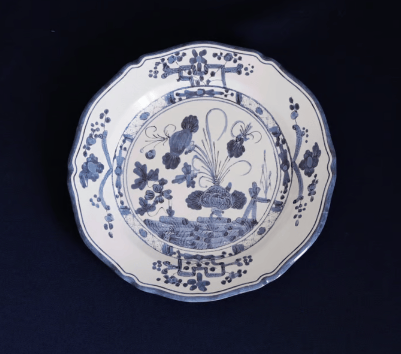 A blue and white melamine plate with a scalloped rim, featuring a detailed blue floral and landscape illustration in the center, set against a dark navy background.