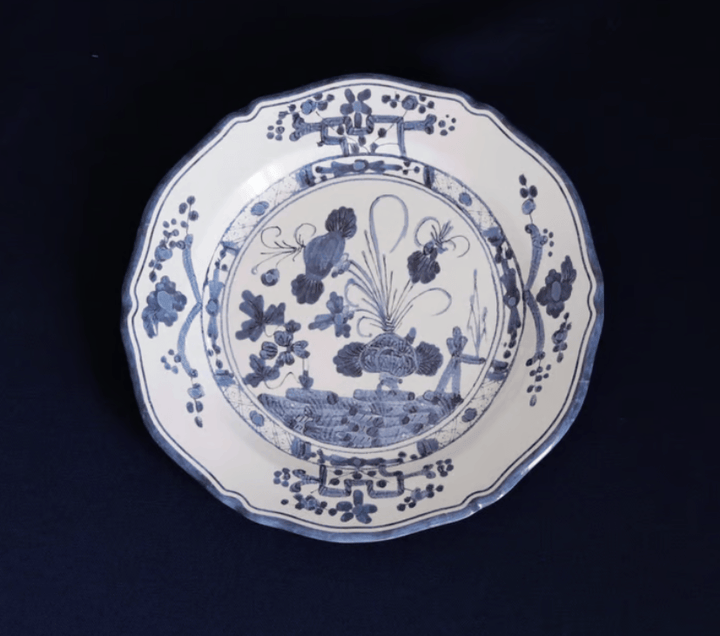 A blue and white melamine plate with a scalloped rim, featuring a detailed blue floral and landscape illustration in the center, set against a dark navy background.