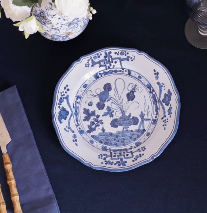A round white melamine plate with blue floral and landscape illustrations, styled on a dark navy tablecloth with a vase of white flowers and a knife partially visible.