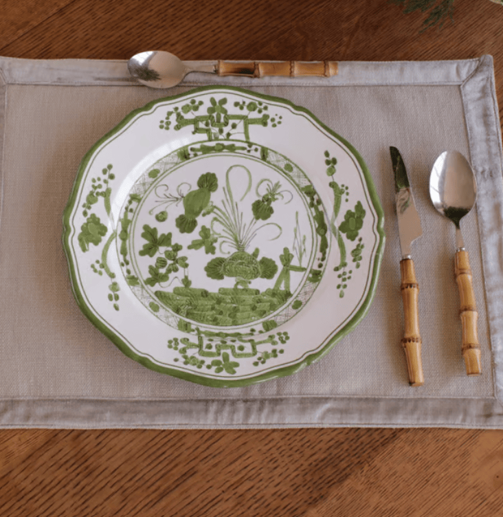 A green and white patterned melamine plate is set on a beige placemat with a bamboo-handled spoon, knife, and spoon on a wooden table.