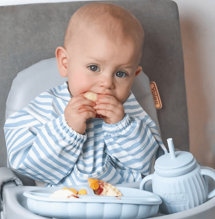 Baby in a high chair eating with a teapot and plate in front
