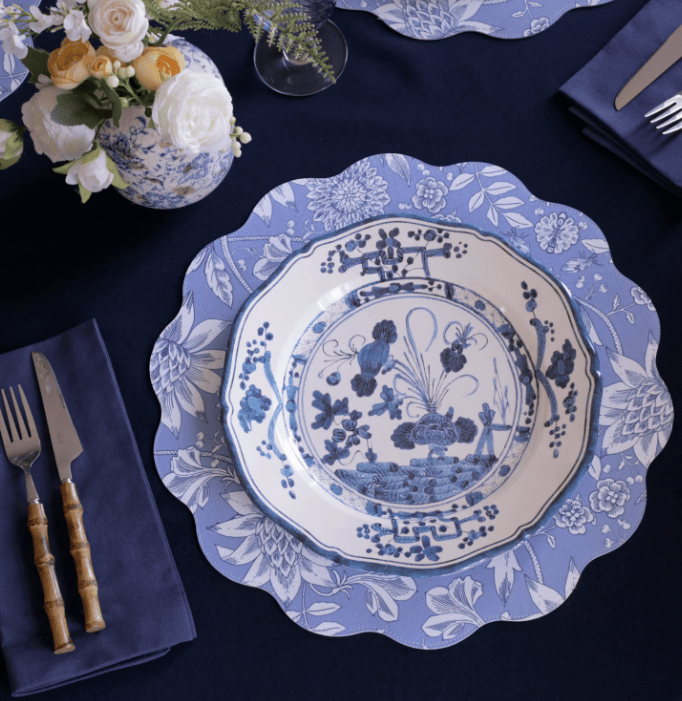 A blue and white melamine dinner plate with a scalloped edge, styled on a dark navy tablecloth with matching napkins, cutlery, and a floral vase.