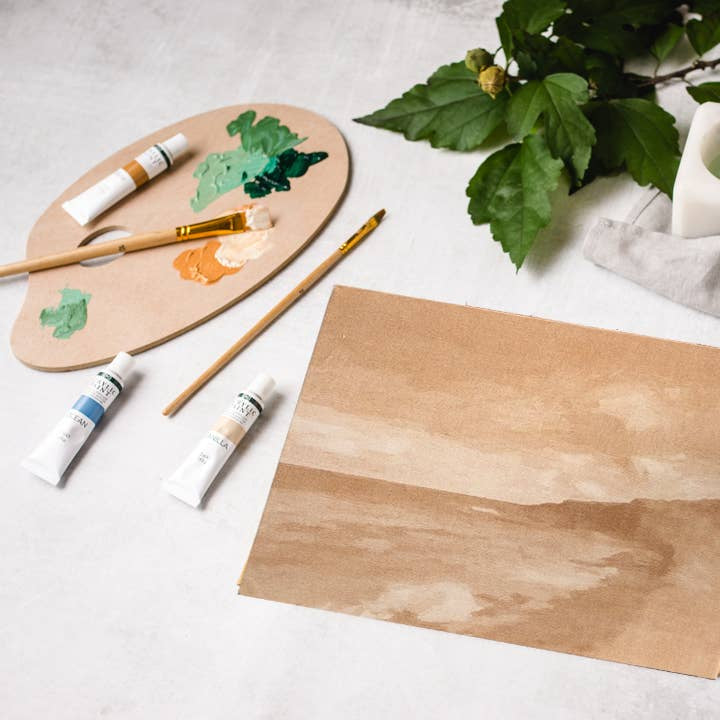 Art supplies including a paint palette, brushes, and a piece of paper on a light surface.