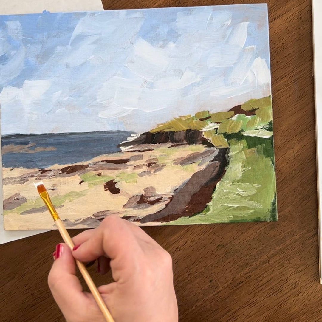 Painting of a coastal scene on a wooden surface with a hand holding a paintbrush.