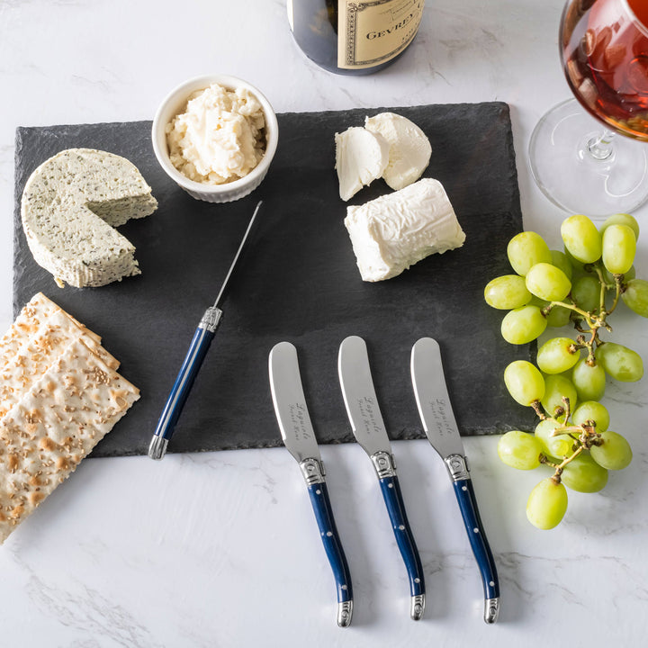 Cheese platter with cheeses, crackers, grapes, and navy blue handled spreaders on a marble surface.