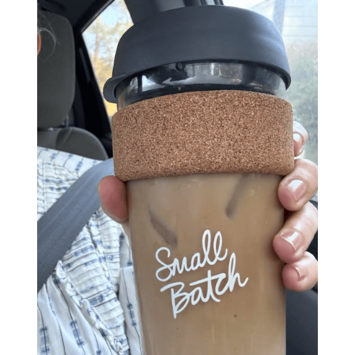Hand holding a coffee cup with a cork sleeve and 'Small Batch' text, inside a vehicle.