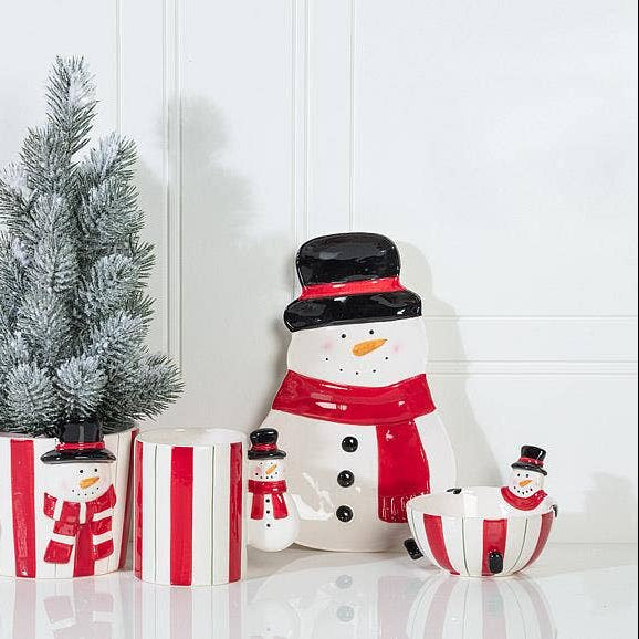 A holiday-themed ceramic set featuring snowman figurines, striped cups, and a small frosted artificial tree on a white surface against a white paneled wall.