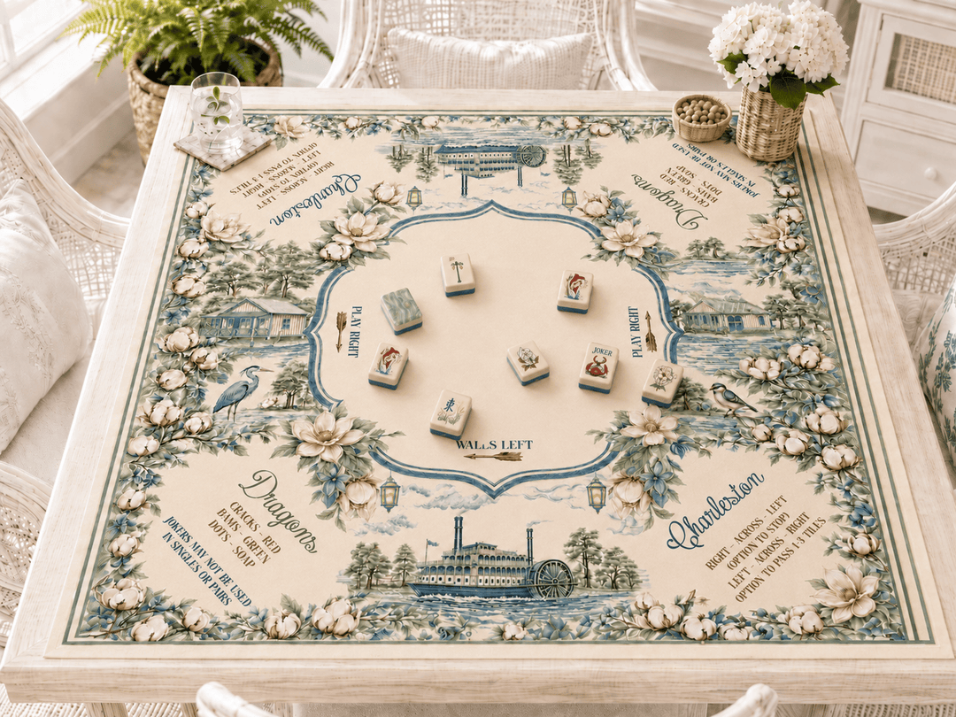 Mahjong mat with floral illustrations and printed text, set on a white wicker table in a bright room with white chairs and flowers.