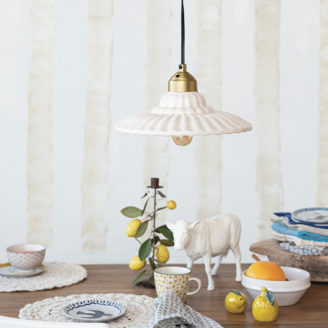 A white ribbed ceramic pendant light with a brass-colored socket and black cord hangs over a styled tabletop scene with a cow figurine, lemons, cups, and plates against a soft cream background.