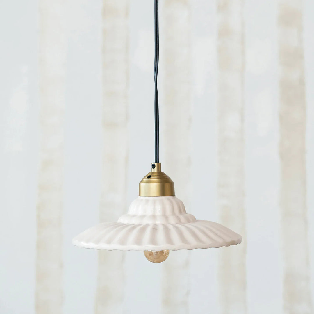 A white ceramic pendant light with a flared, ribbed shade and a brass-colored metal top, suspended by a black cord against a soft, light beige background.