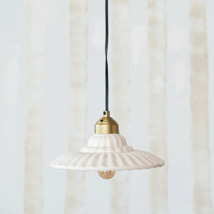 A white ceramic pendant light with a flared, ribbed shade and a brass-colored metal top, suspended by a black cord against a soft, light beige background.