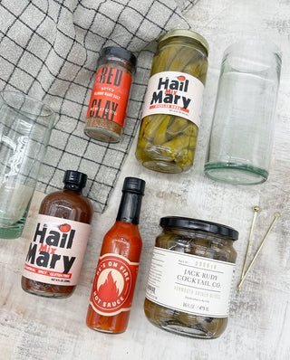 The ultimate gift for the Bloody Mary lover. A reusable hamper is filled to the brim with Bloody Mary essentials: Hail Mary Bloody Mary mix, olives, okra, Bloody Mary salt, fresh hot sauce, two recycled highball glasses, two olive picks and one durable kitchen/bar towel. Carolina Made.