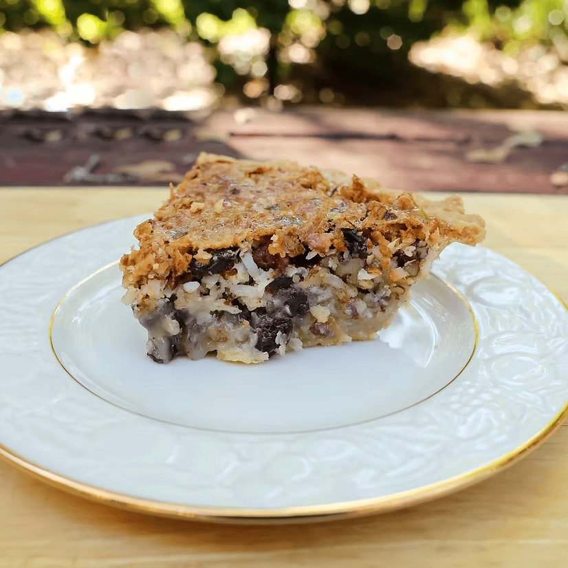 A slice of chocolate chip cookie pie served on a white plate with a gold rim, set on a wooden board with an outdoor garden background.