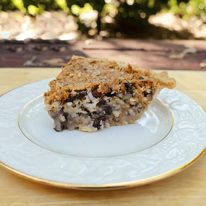 A slice of chocolate chip cookie pie served on a white plate with a gold rim, set on a wooden board with an outdoor garden background.