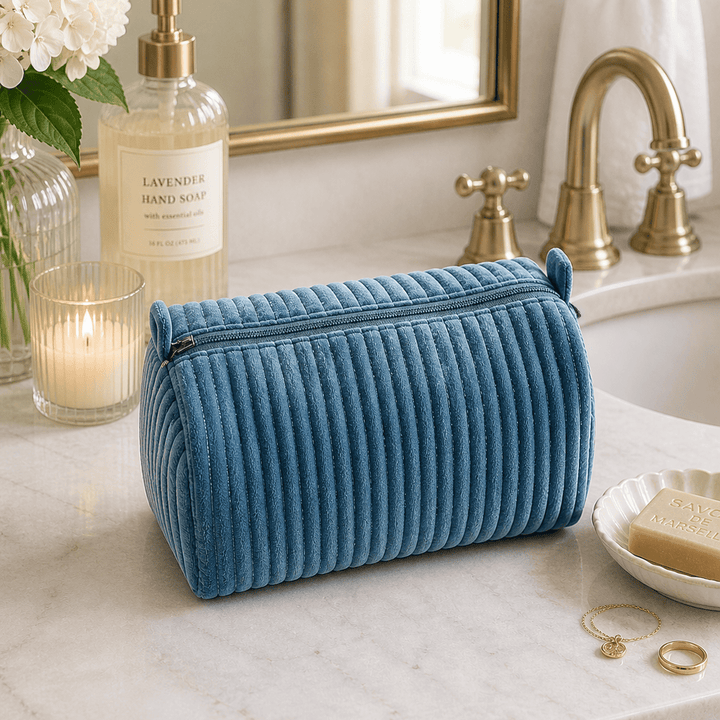 A ribbed blue velvet cosmetic pouch sits on a white marble bathroom counter next to a lit candle, lavender hand soap, and a gold faucet.