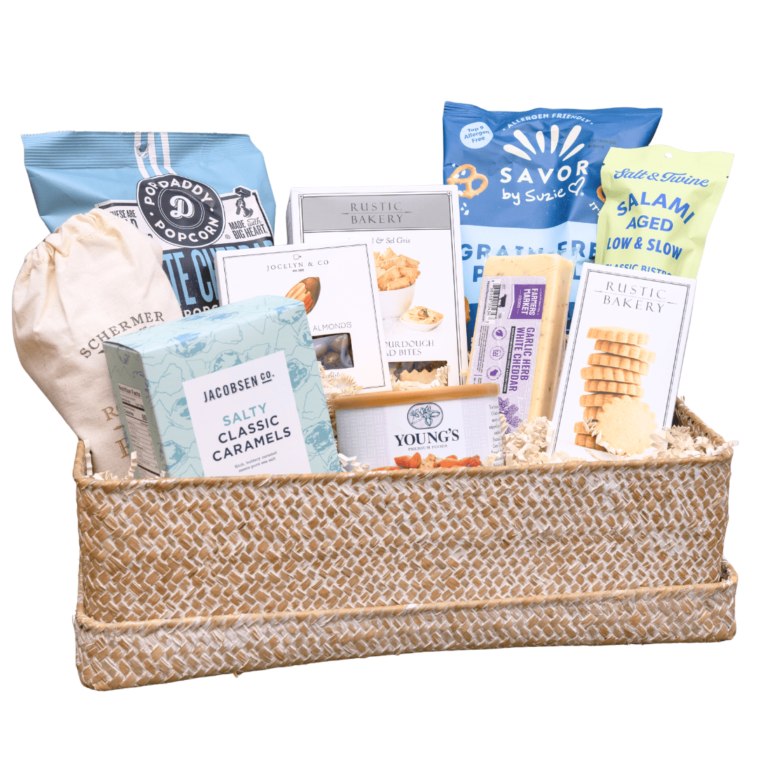 A woven natural-fiber gift basket filled with assorted packaged snacks and baked goods, including bags of chips, crackers, caramels, and pastries, photographed on a plain white background.